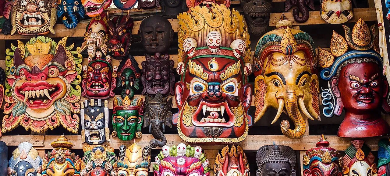 a vibrant collection of intricately carved and painted wooden masks, likely of Nepalese or Tibetan origin, densely arranged and displayed on a surface. The masks vary in size and detail, featuring stylized faces with exaggerated features such as wide eyes, bared teeth, and prominent noses, many adorned with elaborate headpieces and patterns.