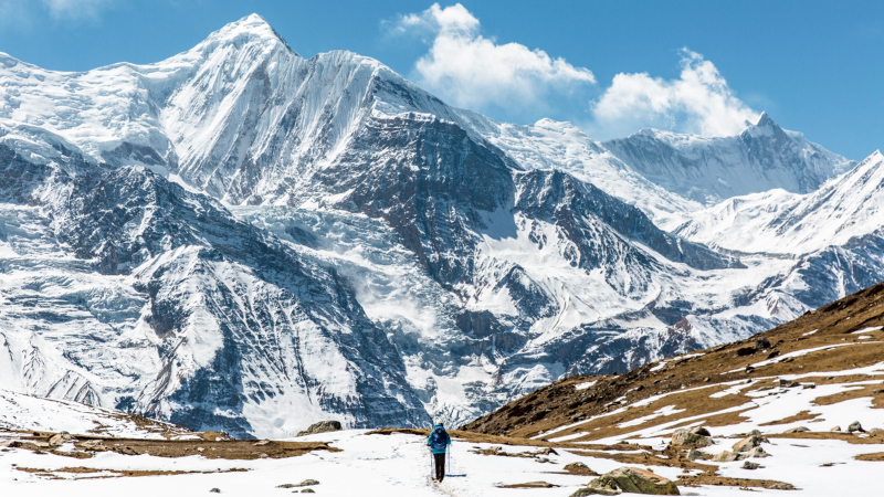Annapurna Circuit solitary figure stands on a rocky, snow-dusted path, gazing up at a majestic, towering snow-capped mountain range under a clear sky