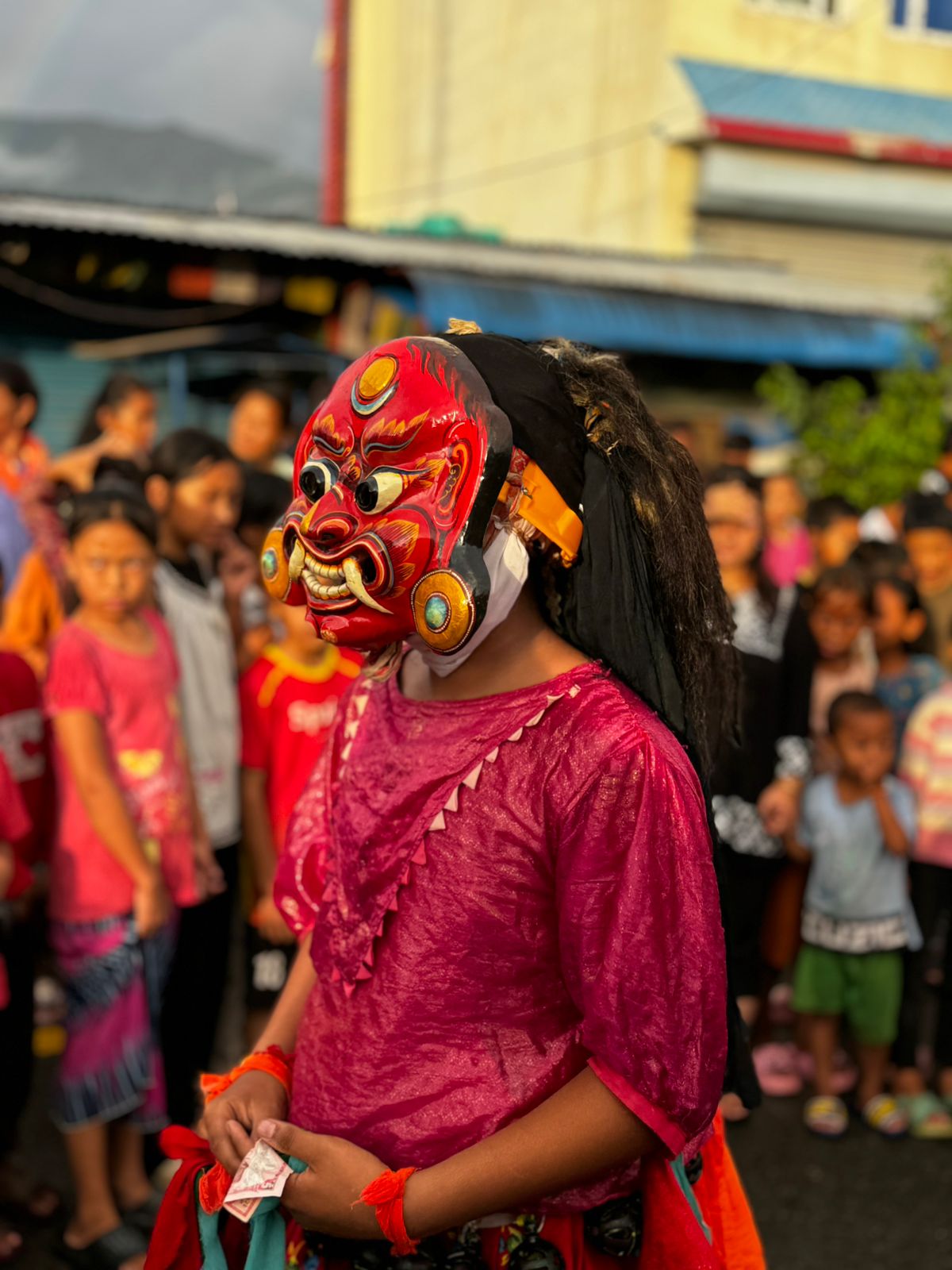 "A person in a red traditional costume and an elaborate, fierce-looking red mask with large ears and black feather-like adornments stands amidst a crowd of people, likely during a festival or cultural event in Nepal, Gai Jatra."