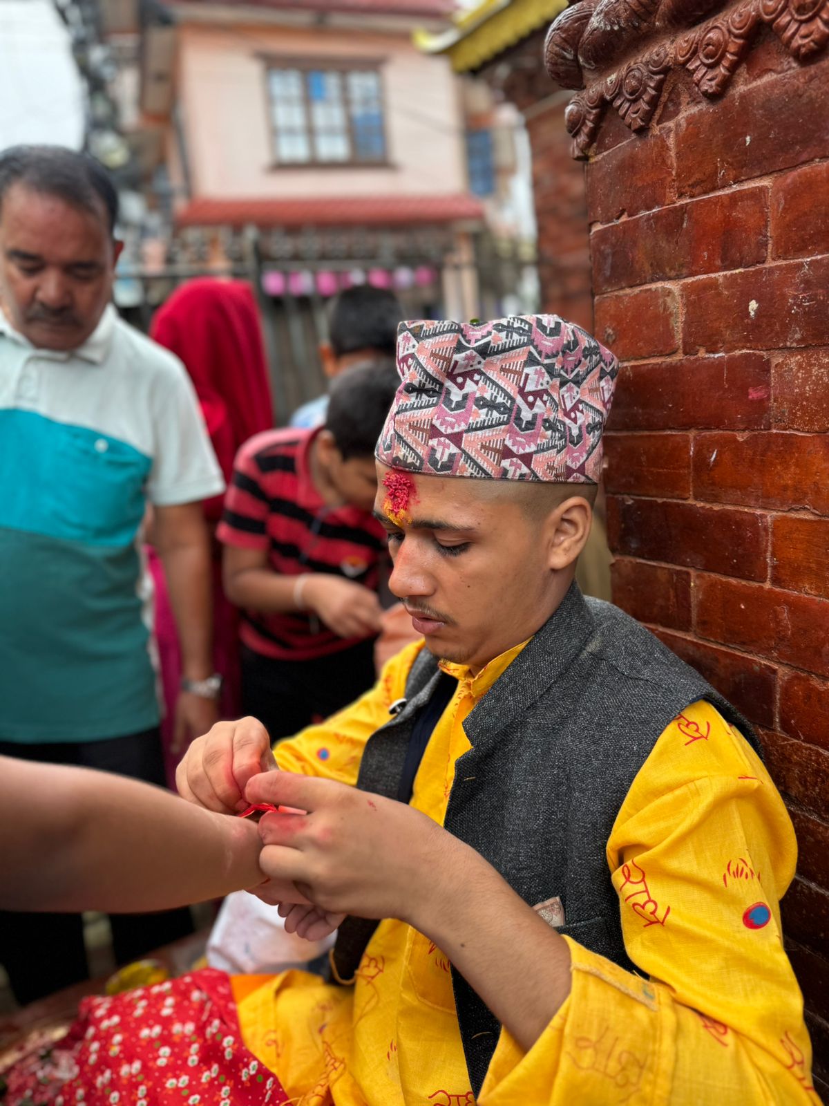 A man in traditional Nepalese attire, including a Dhaka topi and a yellow and black vest over a yellow shirt, is having a sacred thread or bracelet tied around his wrist by another person in a ceremonial setting