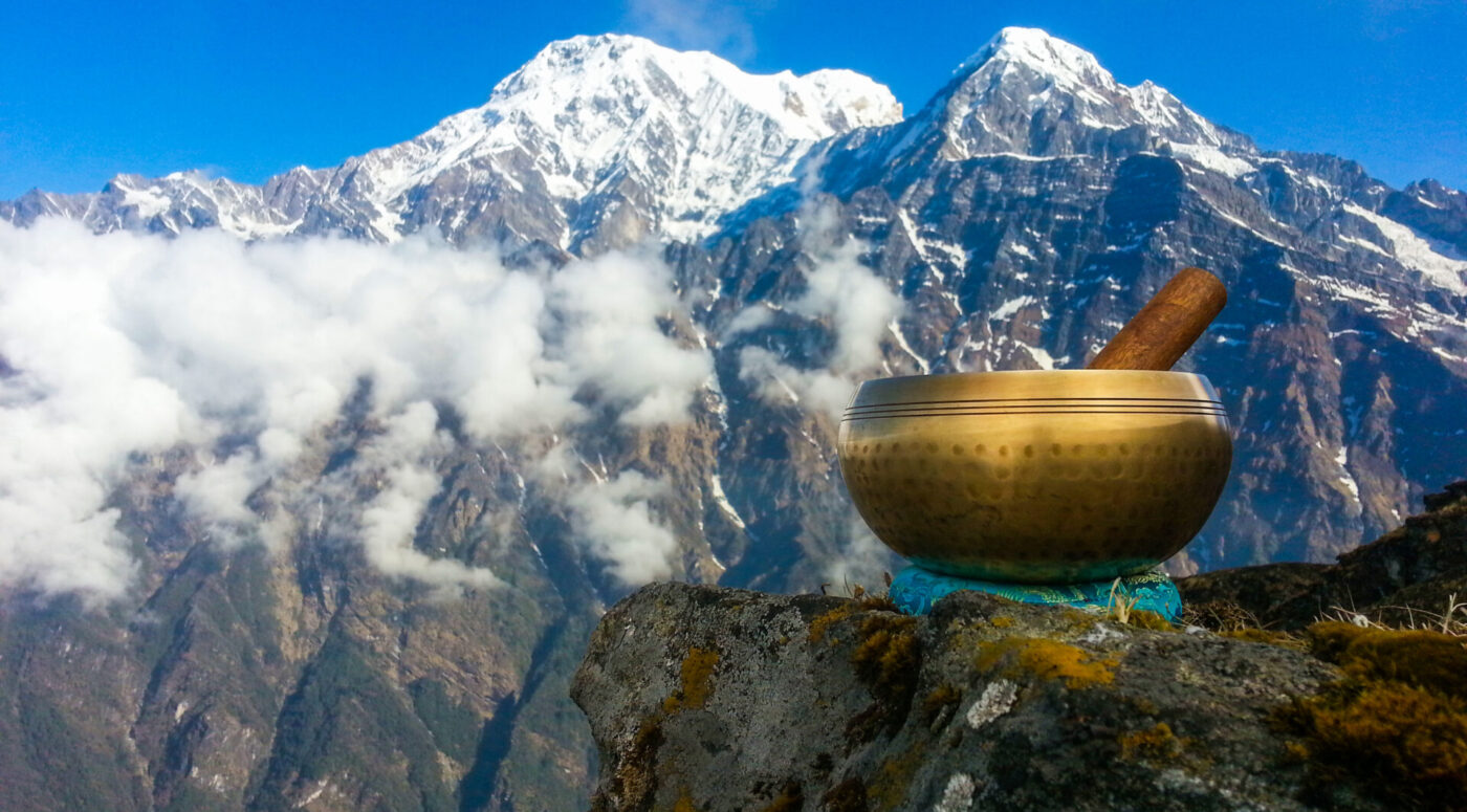 A brass singing bowl with a wooden mallet resting inside, positioned on a rocky outcrop overlooking a majestic mountain range with clouds in the sky, displayed within a digital editing interface