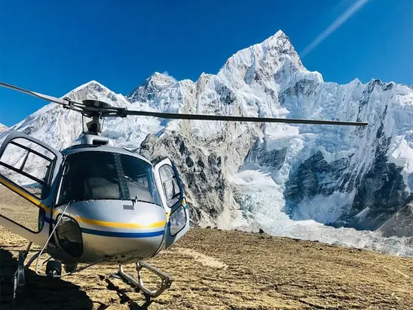 A helicopter with blue and yellow stripes on its side is positioned in the foreground, with its open door visible. In the background, a large, snow-capped mountain range rises under a clear blue sky.