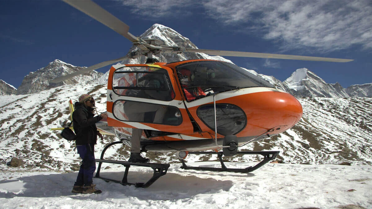 helicopter, orange and white, is positioned on a snow-covered mountain peak with a person standing near its tail rotor, against a clear blue sky.