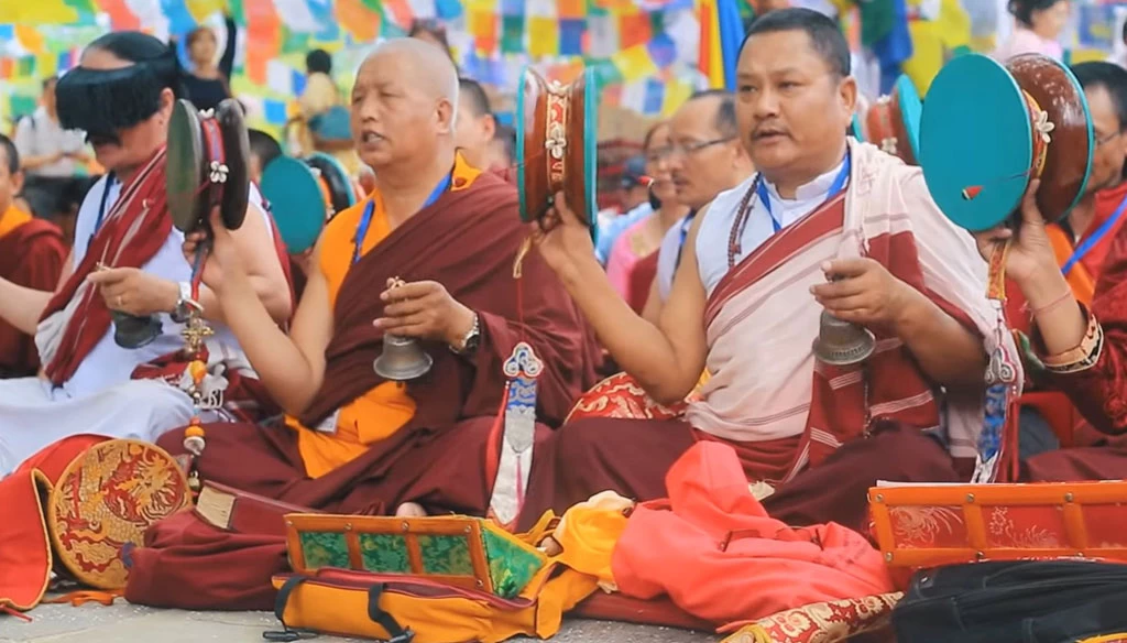 A group of Buddhist monks in robes, holding traditional ceremonial instruments like bells and cymbals.