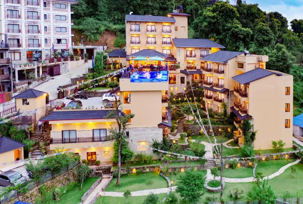 An aerial view of Bar Peepal Resort in Pokhara, Nepal, showcasing its multiple buildings with dark roofs, lush green gardens, winding pathways, and a prominent swimming pool, all illuminated by evening lights