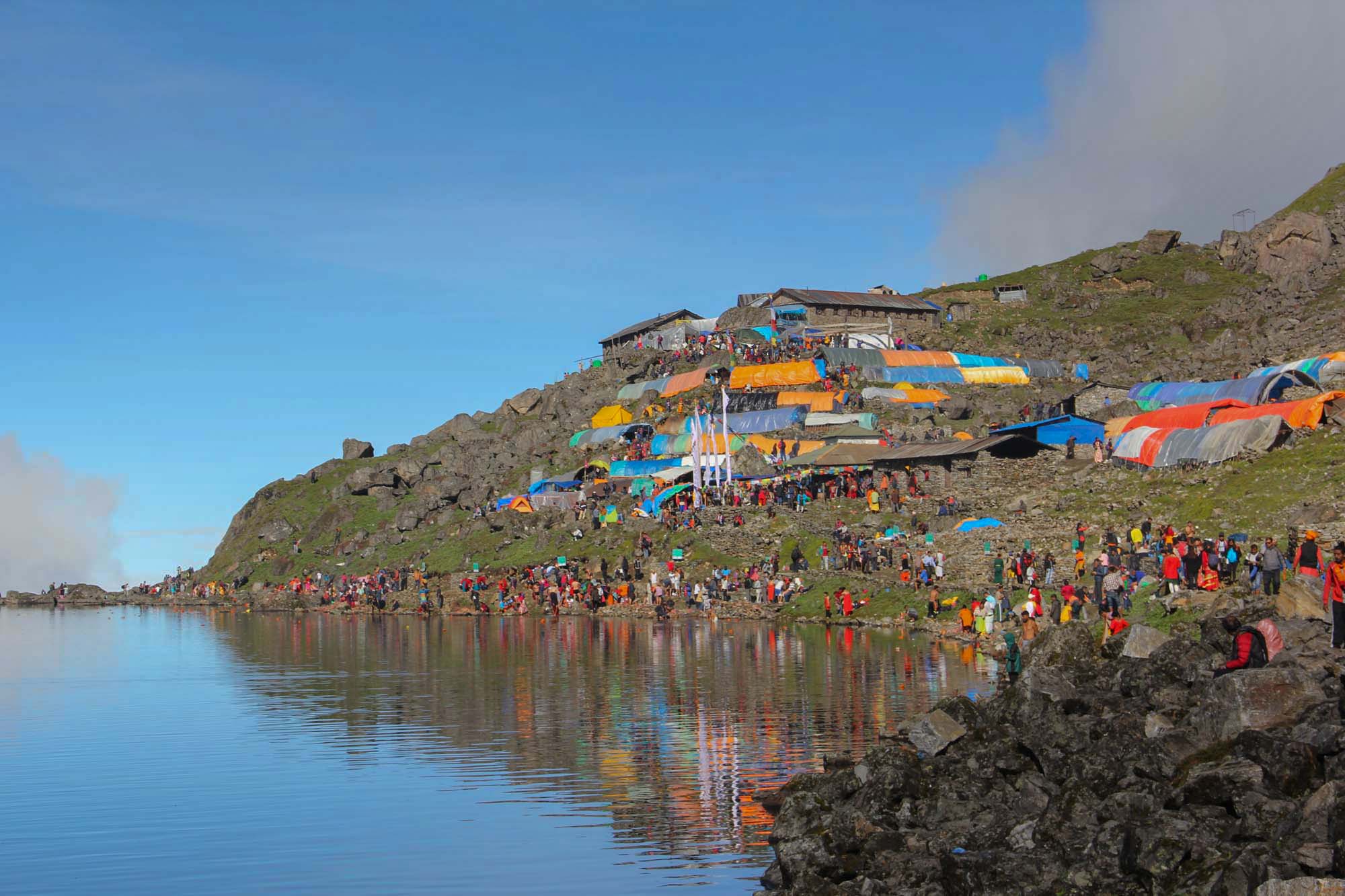 A large crowd of pilgrims and colorful tents line the rocky shore of a clear, high-altitude lake in a mountainous region of Nepal, likely Gosaikunda Lake during a religious festival like Janai Purnima, with reflections shimmering on the water's surface.