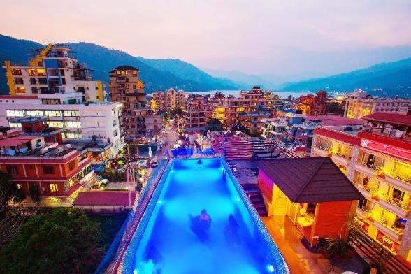 A vibrant, inverted nighttime cityscape with illuminated buildings and a bright blue swimming pool, set against a backdrop of mountains and a twilight sky