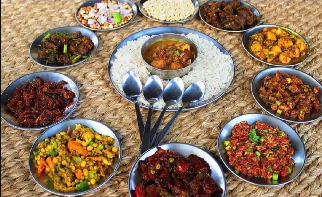 Newari dish called samaya baji is showcases on the picture.dishes arranged on a woven mat. The main focus is a large metal plate containing a mound of white rice and a small bowl of curry. Surrounding this central dish are several smaller metal bowls, each filled with a different type of food, including what appears to be various curries, stir-fried vegetables, and other savory items.