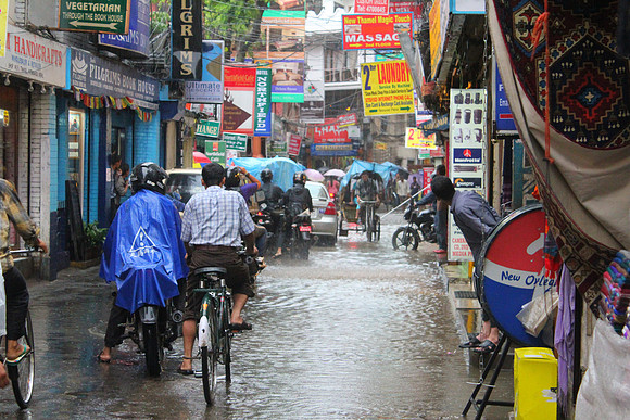 A street scene during rainfall, with multiple individuals on motorcycles and bicycles navigating through flooded areas. The foreground features a person sitting near a colorful barrel