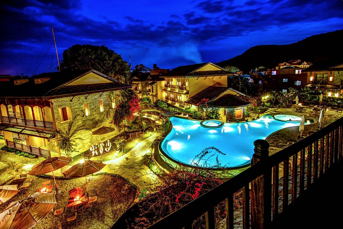 A vibrant, inverted nighttime cityscape with illuminated buildings and a bright blue swimming pool, set against a backdrop of mountains and a twilight sky