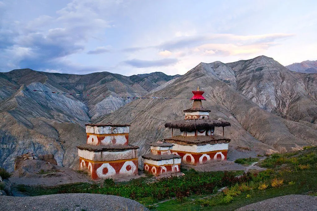 traditional tiered chorten or monastery in the Upper Mustang region of Nepal, built into the side of a rugged, rocky mountain, with distinctive white and red/orange patterns, green foliage nearby, and another smaller structure lower down the slope.