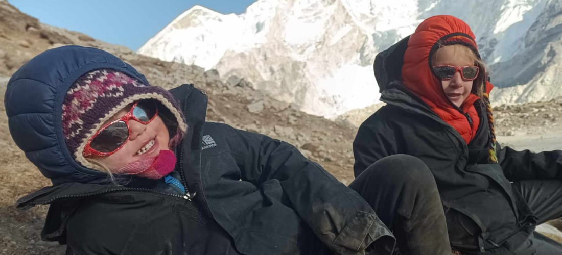 A slightly rotated, medium-close-up shot shows two people bundled in winter gear on a snowy mountain. In the foreground, a person wearing a black hooded jacket, a colorful knit hat, and sunglasses smiles at the camera. In the background, another person, also wearing a hooded jacket and sunglasses, crouches down with a faint smile. Behind them, the jagged, snow-covered peak of a mountain is visible.
