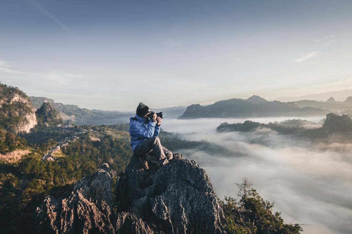 man taking picture while sitting on a rock and surrrounded by hills and clouds