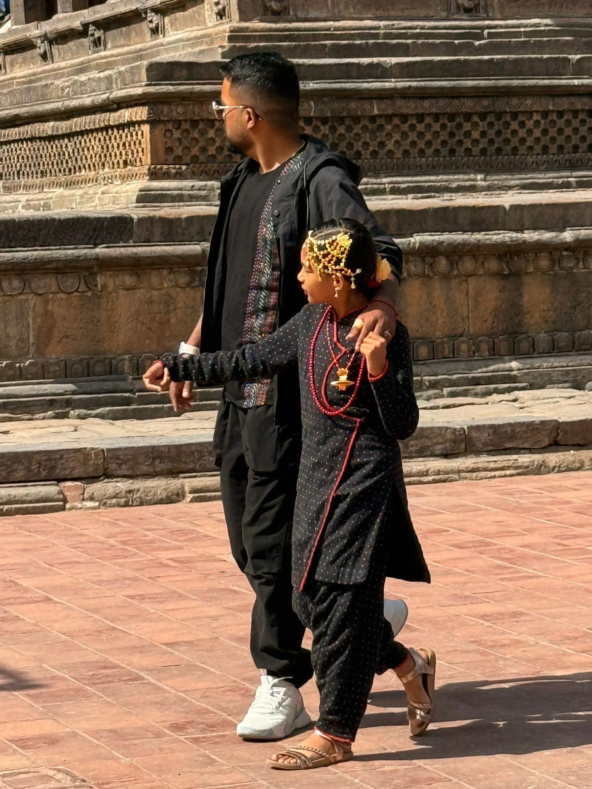 A man wearing a black jacket and sunglasses stands next to a young girl in a long, black traditional dress with red trim, embellished with gold head jewelry and a red beaded necklace. They are walking across a reddish-brown brick plaza.