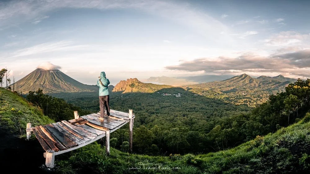 man wearing brown pant and green jacket and clicking pictured of hills while surrounded by greenery