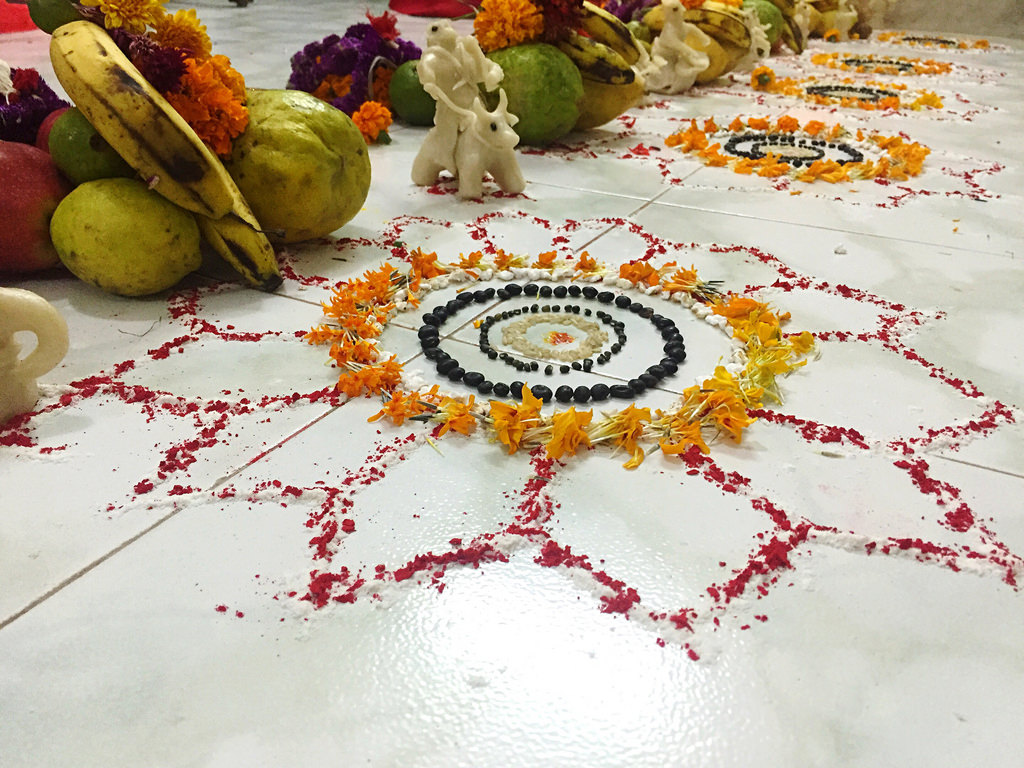 a picture of a rangoli or mandala on a white floor. The decoration is made with red powder, yellow marigold flowers, and black seeds. In the background of the picture on the screen, there are fruits, including guavas and bananas, and more marigold flowers.