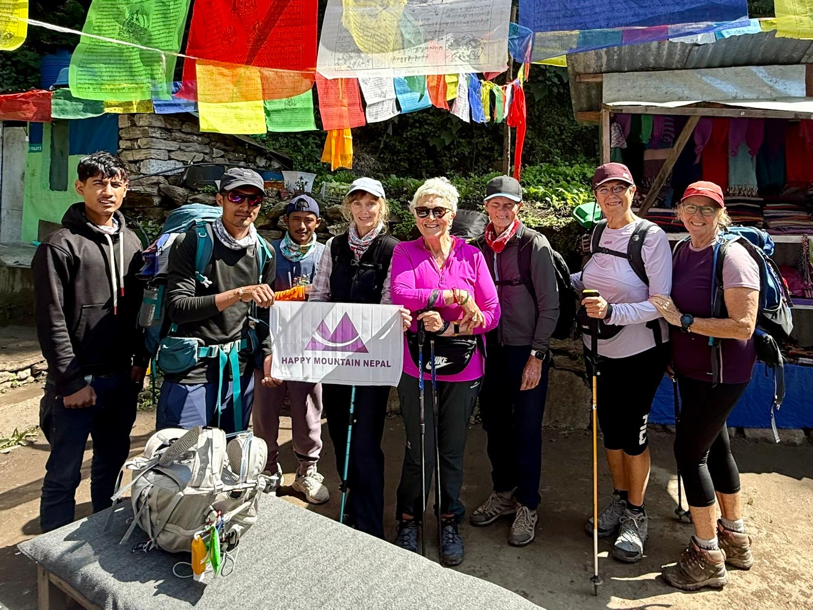 A group of people, including five tourists and several local guides, stands together outside a small shop with colorful prayer flags and goods hanging overhead. The tourists are wearing hiking gear and holding trekking poles. The group is holding a white banner that reads "HAPPY MOUNTAIN NEPAL" with a purple logo.