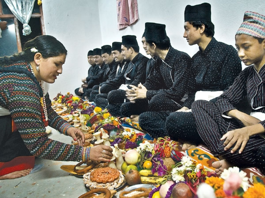 Mha puja  ceremony where several people, some in traditional clothing, are sitting on the floor around a large arrangement of food offerings and other items. The person in the foreground is reaching for something in the arrangement.