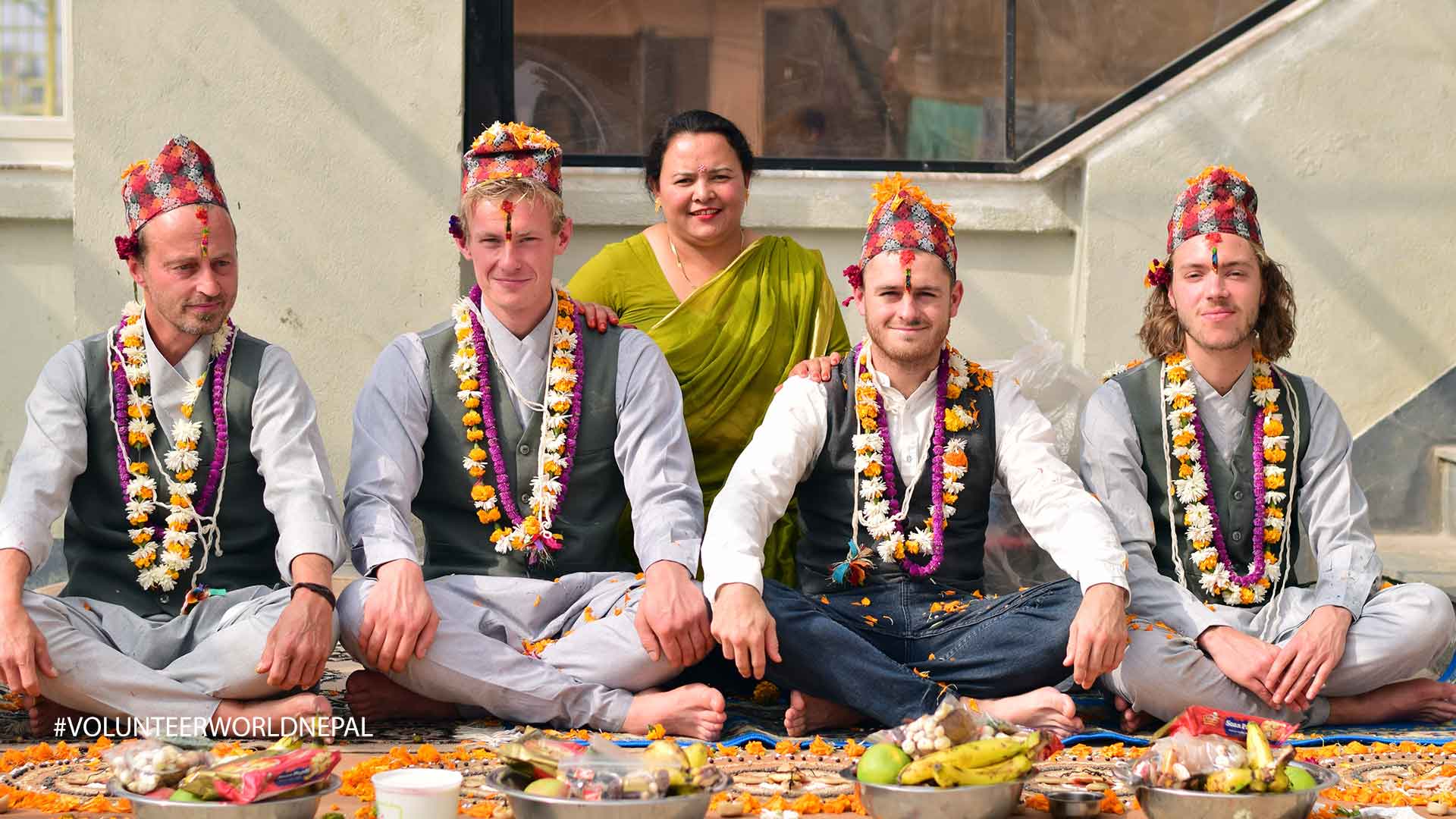 An image of five people, four men and one woman, sitting on the floor during what appears to be a religious ceremony. They are all wearing garlands of flowers and traditional hats. The woman is wearing a green sari during bhaitika in Nepal