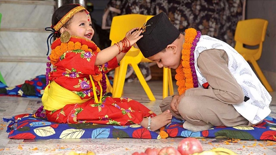 young boy and girl in traditional celebrating Bhaitika.Nepali attire, both wearing marigold garlands. The girl, in a red and orange dress, is smiling and has her hand on the boy's head. The boy is wearing a white vest, brown pants, and a black Dhaka Topi.