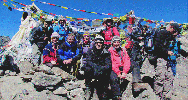 The image shows a group of people posing for a photo on a mountain peak, identified by text on a small flag