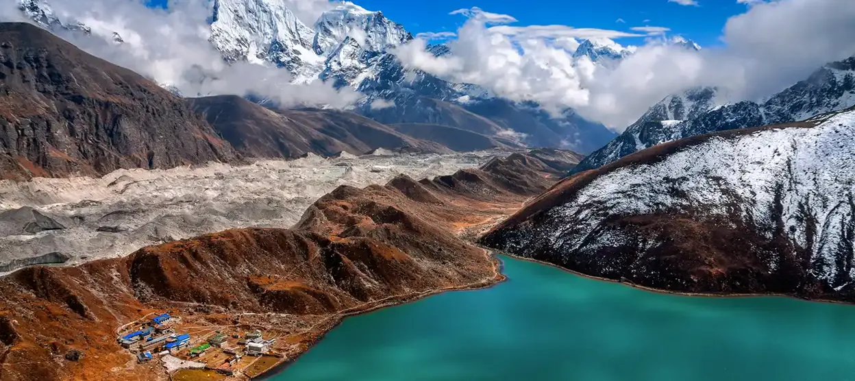 vertical photograph of a high-altitude mountain landscape, likely in the Himalayas, featuring a turquoise lake and a small settlement. It appears to show a popular trekking destination, potentially around the Annapurna region of Nepal.