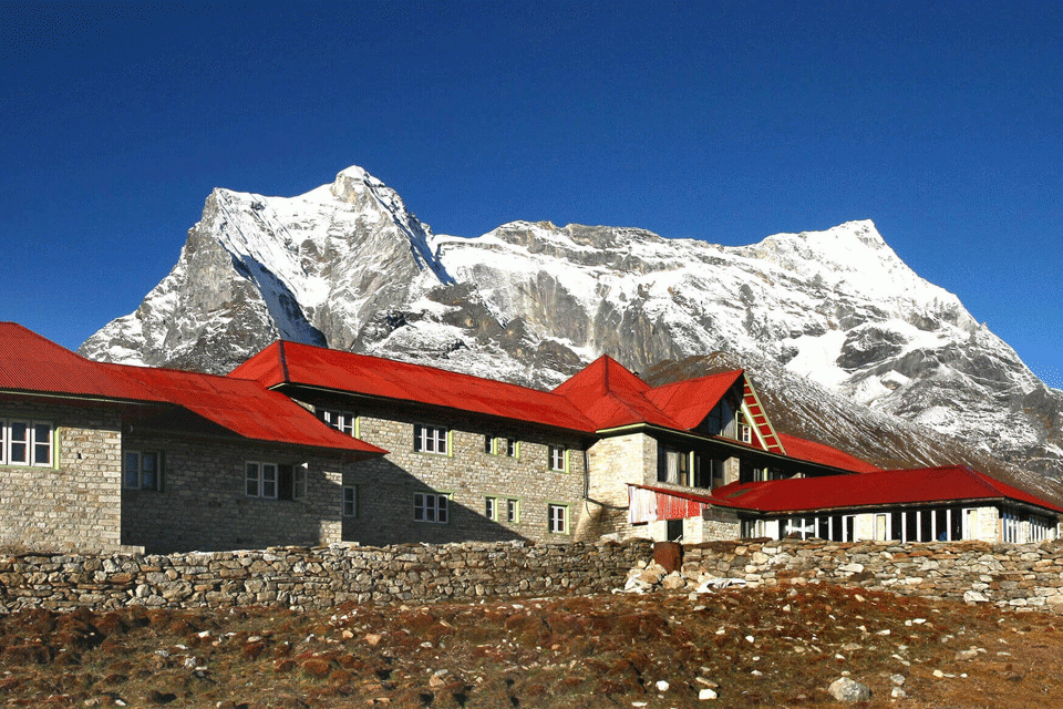 a stone building, likely a tea house or lodge, in a high-altitude, mountainous region, typical of the kongde trek. The visible snow-capped peak suggests an altitude of several thousand meters above sea level.